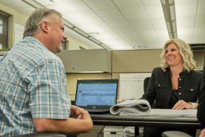 Two coworkers sit across from each other at a desk in an office, a man in a blue plaid shirt facing a smiling woman in a black jacket with a laptop and papers between them, suggesting a discussion over work documents.