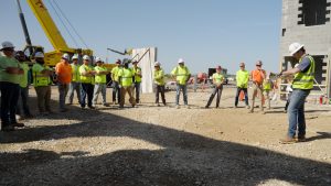 Construction crew in neon vests and hard hats stand in a circle on a dirt site as a supervisor speaks near a concrete wall and crane in the background.